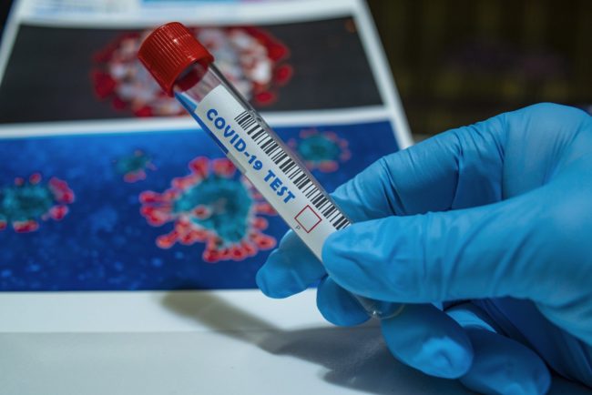 Photo of a hand wearing a blue surgical glove, holding a test tube labeled "COVID-19 Test" in front of photos of magnified viruses.