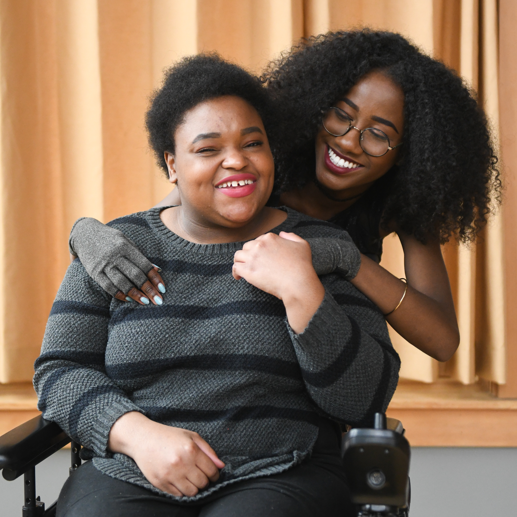 Two young women smiling. One stands behind the other with her arms wrapped around her, seated in her wheelchair. 