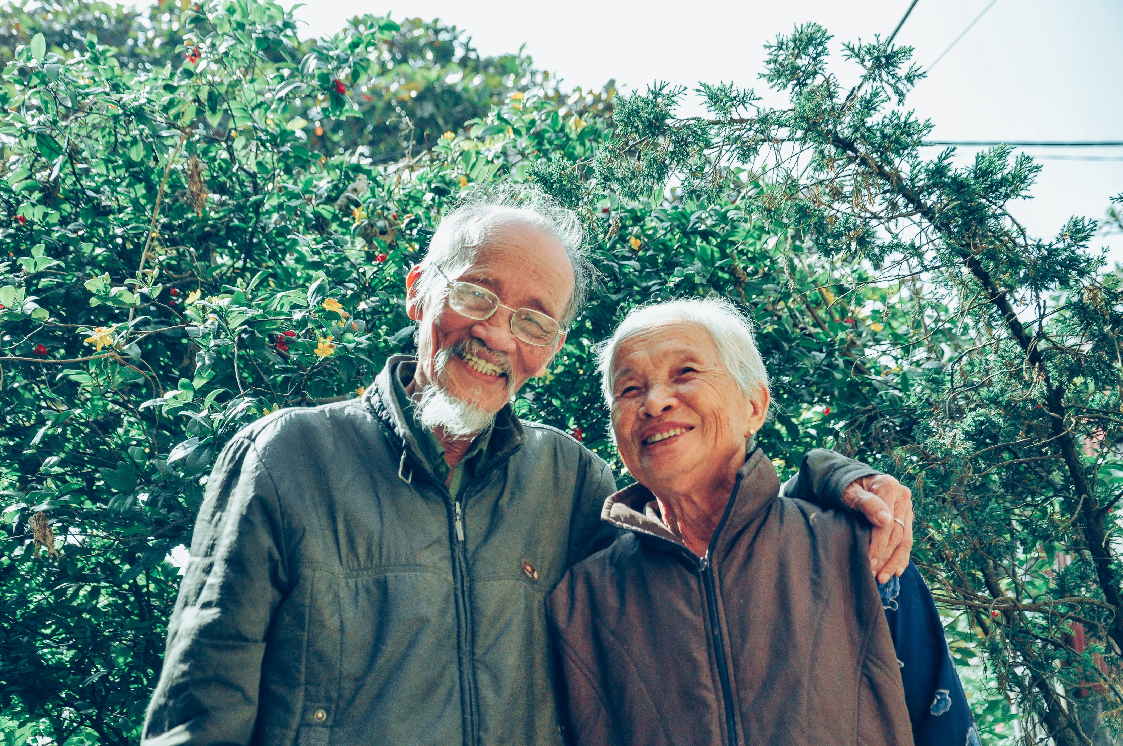 Senior couple smiling, the man's arm is on the woman's shoulder as they pose outdoors. 