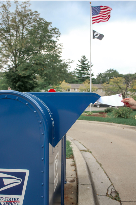 A person's hand is shown placing a ballot into a mailbox, under an American flag, along a street that runs in front of a row of houses