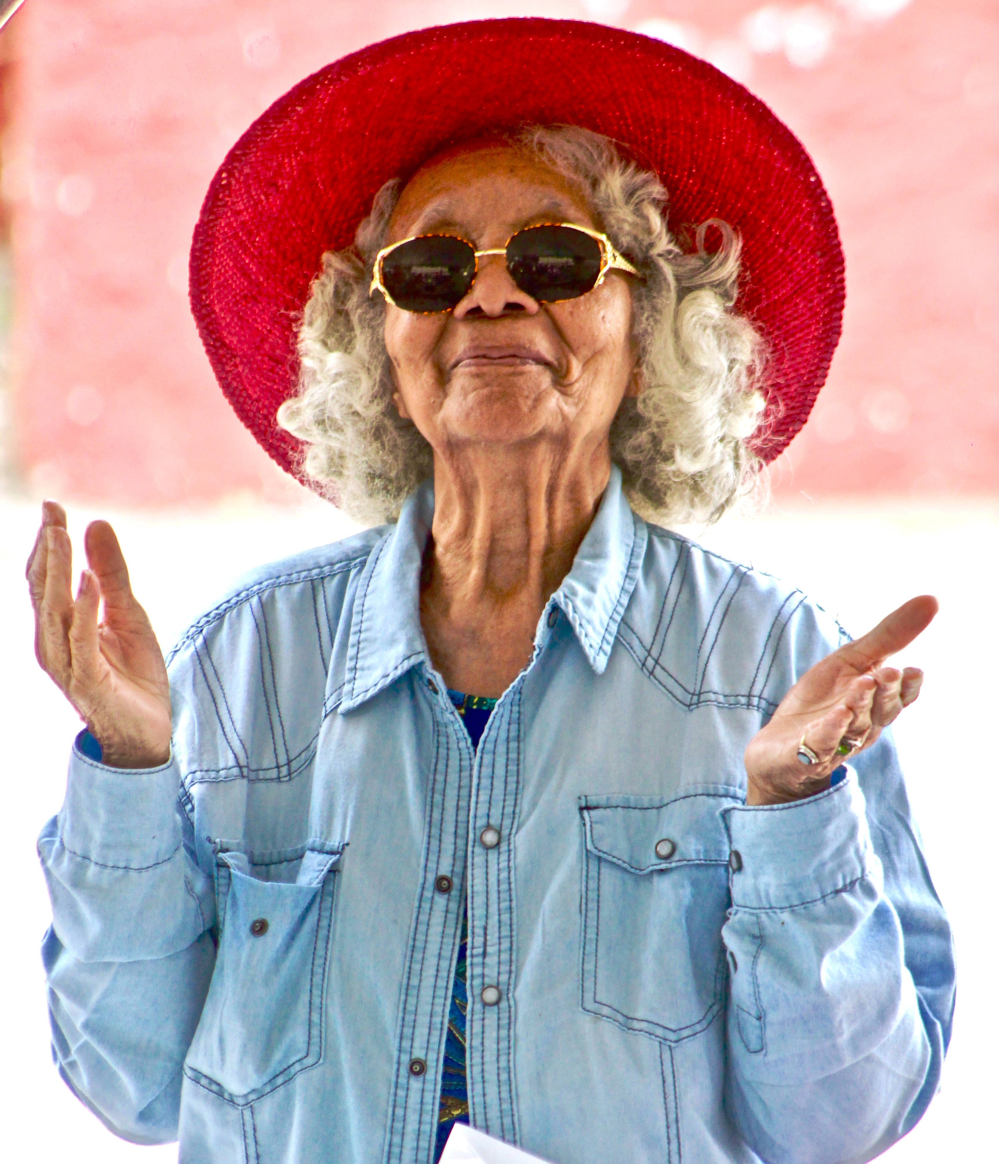 Senior woman smiling, wearing a red, wide brimmed sun hat, sunglasses, and denim shirt. 