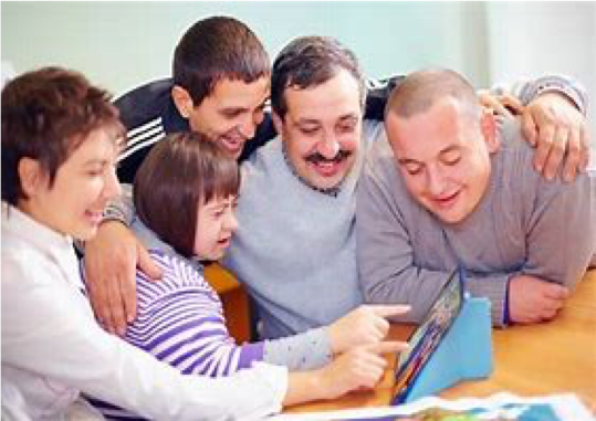 Picture of four family members huddled arm in arm around young woman with developmental disabilities. They are looking at a monitor, and a woman seated behind her is pointing at the screen along with her. 
