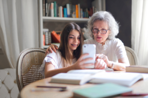 An older white woman seated at a study table with her smiling granddaughter, who is showing her something on her mobile phone