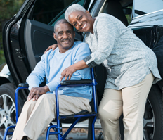 A older, African-American couple together, they are both smiling. The woman has her arm around her partner who uses a wheelchair