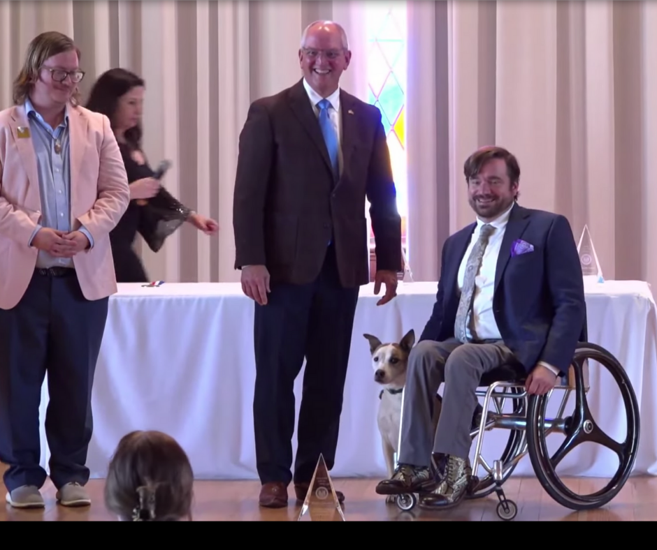 Governor Jon Bel Edwards standing next to Phoebe, a white and tan mixed breed service dog, and Jonathan, a young white man with short brown hair, moustache and beard, wearing a suit and tie with purple handkerchief fold in his breast pocket. They are smiling for a picture as they accept the award. They are flanked by presenter Chris Turner Neal, a white man with long dark blonde hair, wearing a pink blazer. Bambi Polotzola, a white woman with dark hair, wearing a black dress, can be see carrying a microphone from behind a display table covered by a white tablecloth onstage.