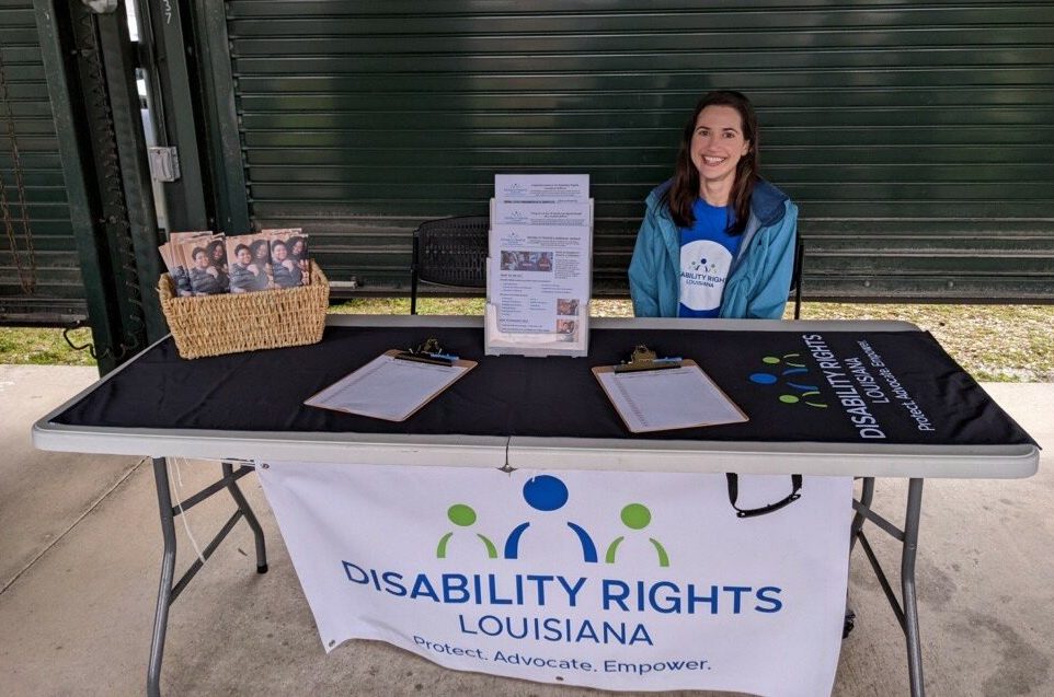 Photo of DRLA attorney, Shannon Barnes, a white woman with dark hair, smiling and sitting behind a table containing flyers, brochures, and fronted by a banner with the Disability Rights Louisiana logo on it in blue and green against a white background