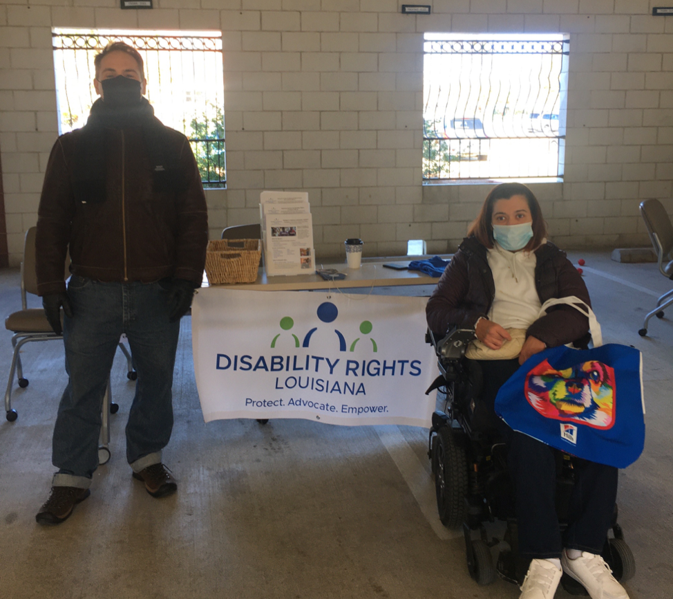 Photo of DRLA Director of Policy & Community Engagement, Tory Rocca and Policy Analyst, Ashley Volion, stand to each side of a table with a banner displaying the DRLA logo in blue and green against a white background. The table has a display of flyers and other items, housed in a garage with open windows displayed in the background
