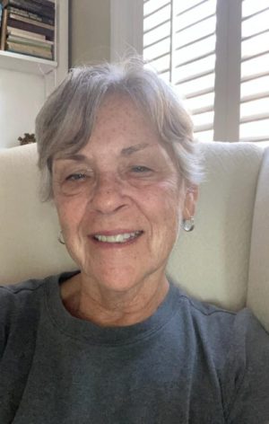 Photo of Lois V. Simpson, a white woman with grey hair wearing a blue shirt, sitting on a white sofa and smiling